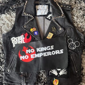 May include: A black denim vest with silver studs along the collar and shoulders. Patches include "REBEL", "NO KINGS NO EMPERORS", and a Mickey Mouse-inspired design. Zippers and various pins add detail to this punk-inspired garment.