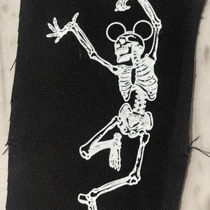 May include: A white skeleton with Mickey Mouse ears is dancing on a black fabric patch. The skeleton is in a dynamic pose with arms raised and legs spread.