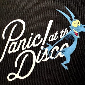 May include: Black background with the white text "Panic! at the Disco" in a cursive font. A blue cartoon pterodactyl with yellow eyes and a red mouth is on the right side of the image.