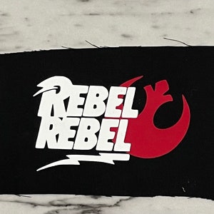 May include: Black rectangular patch with the words "REBEL REBEL" in white, stacked vertically. A red symbol is on the right. The patch is on a white and gray marble surface.