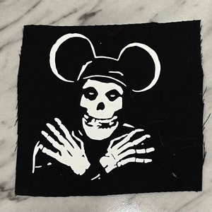 May include: Black square patch featuring a white skull wearing a hat with mouse ears, crossed skeletal arms, and a wide grin. The patch has a raw edge and is likely for sewing or ironing onto fabric. The design is a parody of a famous cartoon character.