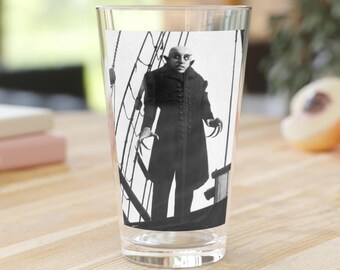 May include: A clear pint glass featuring a black and white image of a vampire character from a classic horror film. The image shows the vampire standing on a ship deck. The glass has the text "MYSTERY SUPPLY G" printed on it.
