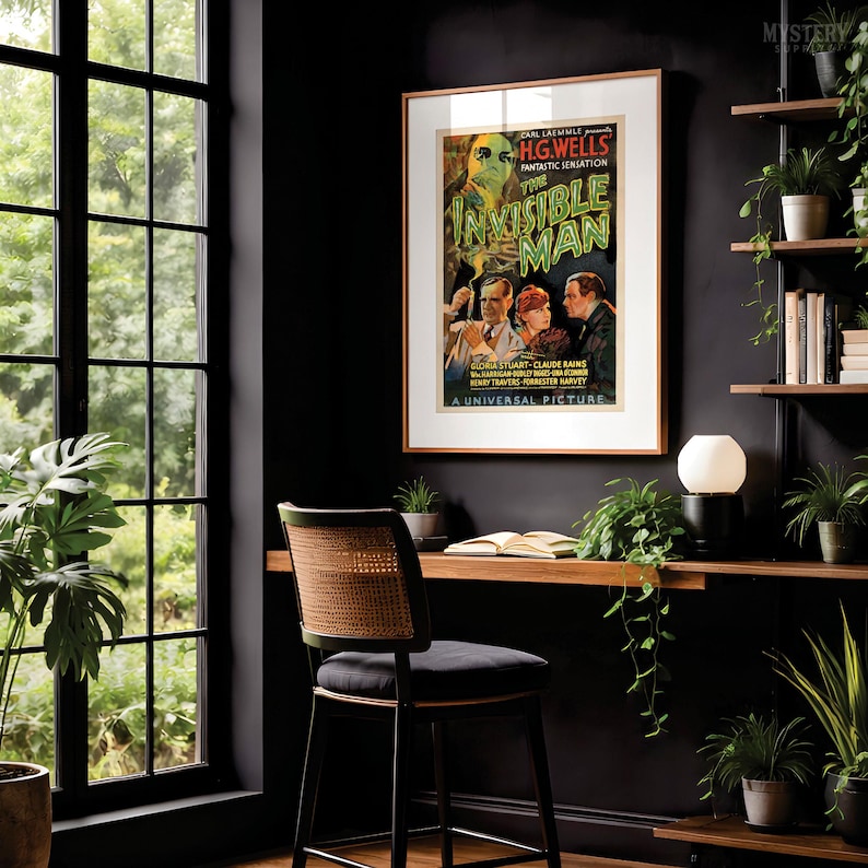 May include: A framed movie poster for The Invisible Man by H.G. Wells, featuring a green-tinted figure and three actors. The poster is displayed on a dark wall above a wooden desk with a chair, a lamp, and several potted green plants.