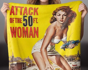 May include: A yellow throw blanket with the text "Attack of the 50 ft. Woman" in red and black letters. The image shows a woman in a white bikini top and skirt standing on a highway overpass, with a car in her hand.