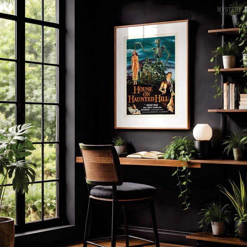 May include: A framed movie poster for House on Haunted Hill featuring a skeleton, a woman in a dress, and a man in a suit. The poster is displayed on a dark wall above a wooden desk with a chair, a lamp, and several potted green plants.