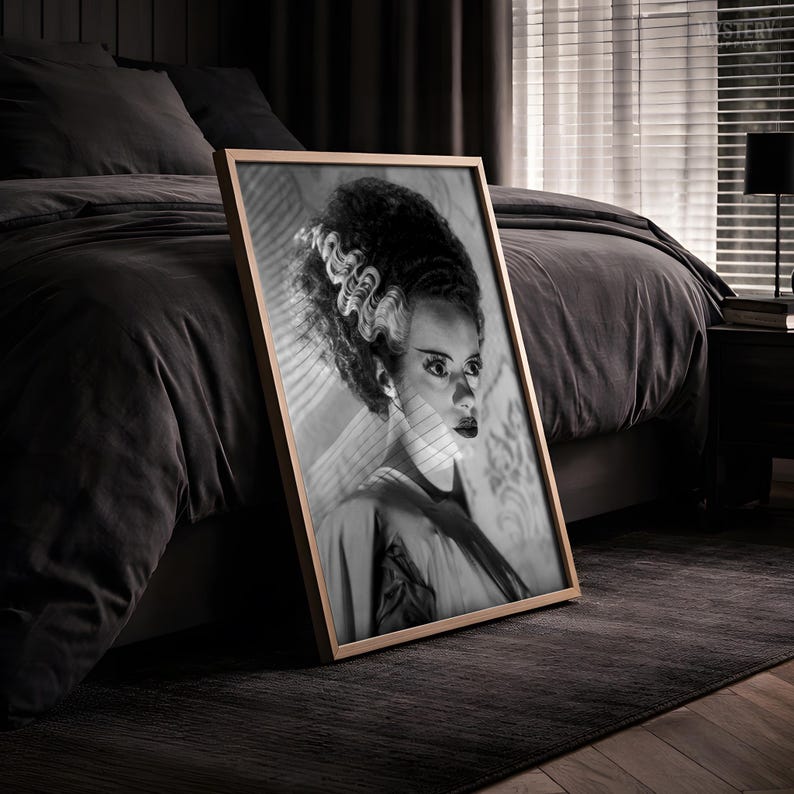 May include: A framed black and white portrait print featuring the Bride of Frankenstein with her signature tall, wavy hair with light streaks, dark eye makeup, and a draped garment, presented in a simple, thin, light-colored wooden frame.