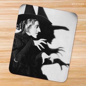 May include: A black and white photo of a woman dressed as a witch with a pointed hat and long, sharp fingernails. Her shadow is cast on the wall behind her, and it also has long, sharp fingernails.