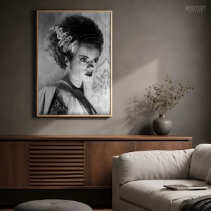May include: A black and white framed wall art print featuring a portrait of the Bride of Frankenstein with her signature tall, wavy hair with white streaks, dark eyes, and dark lips, set against a patterned wallpaper background.