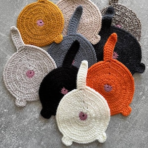 Può includere: Un set di nove sottopentola in crochet a forma di gatto. I sottopentola sono di vari colori, tra cui bianco, grigio, arancione e nero. Ogni sottopentola ha un bottone rosa cucito al centro.