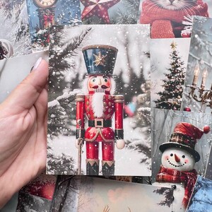 Peut inclure: Une carte postale rectangulaire représentant un casse-noisette central en uniforme rouge et noir avec un grand chapeau, debout dans une forêt enneigée. L'image est entourée d'autres cartes sur le thème des fêtes montrant un chat, un bonhomme de neige et un cerf.