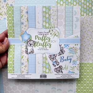 Pode incluir: Um conjunto de 10 folhas de papel de dupla face medindo 20 cm por 20 cm, intitulado Puffy Fluffy Boy da Fabrika Decoru. A coleção apresenta ilustrações de coalas, estrelas azuis e padrões de gotas, laços e grades em tons de verde e azul.