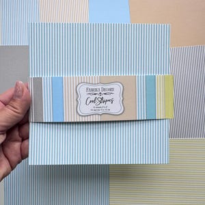 Könnte beinhalten: Ein Set aus zehn 15 x 15 cm großen Papierbögen mit dem Titel Fabrika Decoru Cool Stripes. Das Papier hat vertikale hellblaue und weiße Streifen. Eine Banderole zeigt den Markennamen und den Titel 10 sheets 15 x 15 cm.