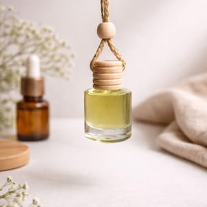 May include: A small, clear glass bottle filled with yellow liquid, suspended by a natural rope and wooden bead. The bottle has a wooden cap. A brown glass bottle and a wooden coaster are in the background. The image has a soft, natural aesthetic.