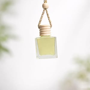 Op de afbeelding: Een kleine, vierkante glazen fles gevuld met een lichtgele vloeistof, opgehangen aan een gevlochten beige koord en een houten kraal. De fles heeft een lichtgekleurde houten dop. De achtergrond is zacht en wazig.