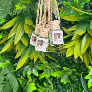 Puede incluir: Tres pequeños frascos de vidrio con tapas de madera cuelgan de una cuerda trenzada. Cada frasco tiene una etiqueta con una ilustración de dibujos animados de perros. Los frascos están rodeados de follaje verde y amarillo.