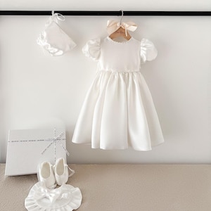 Pode incluir: Vestido de beb&eacute; de cetim branco com mangas bufantes, um gorro de cetim branco e sapatos de cetim branco. O vestido est&aacute; pendurado em um cabide de madeira com uma fita rosa. Os sapatos est&atilde;o em uma caixa branca com uma fita branca.