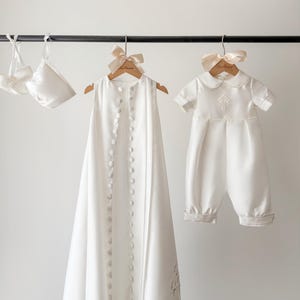 Peut inclure: Ensemble de vêtements de baptême blancs suspendus à une barre noire. L'ensemble comprend une longue robe boutonnée, un barboteuse à manches courtes avec une croix et une paire de chaussons. Chaque article est sur un cintre en bois avec un nœud.
