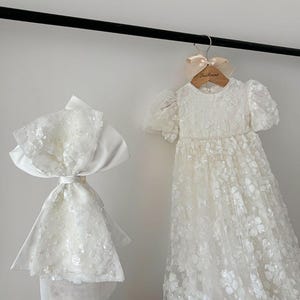 May include: A white, sequined, tulle dress with a satin sash and a matching white, sequined, tulle bonnet. The dress is hanging on a wooden hanger with a pink ribbon and the text "La Reine" printed on it.