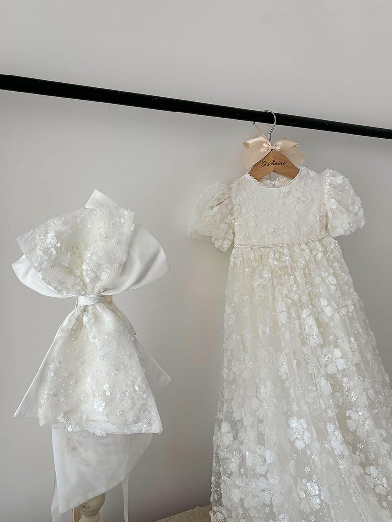 May include: A white, sequined, tulle dress with a satin sash and a matching white, sequined, tulle bonnet. The dress is hanging on a wooden hanger with a pink ribbon and the text "La Reine" printed on it.
