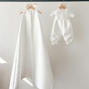 Op de afbeelding: Twee witte satijnen dooppakjes hangen aan hangers. Het pakje aan de linkerkant is een lange jurk met een rij knopen aan de voorkant. Het pakje aan de rechterkant is een romper met korte mouwen en een kraag.