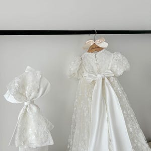 May include: White christening gown with a satin sash and a matching bonnet. The gown is decorated with delicate floral embroidery and sequins.