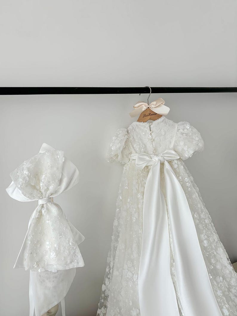 May include: White christening gown with a satin sash and a matching bonnet. The gown is decorated with delicate floral embroidery and sequins.