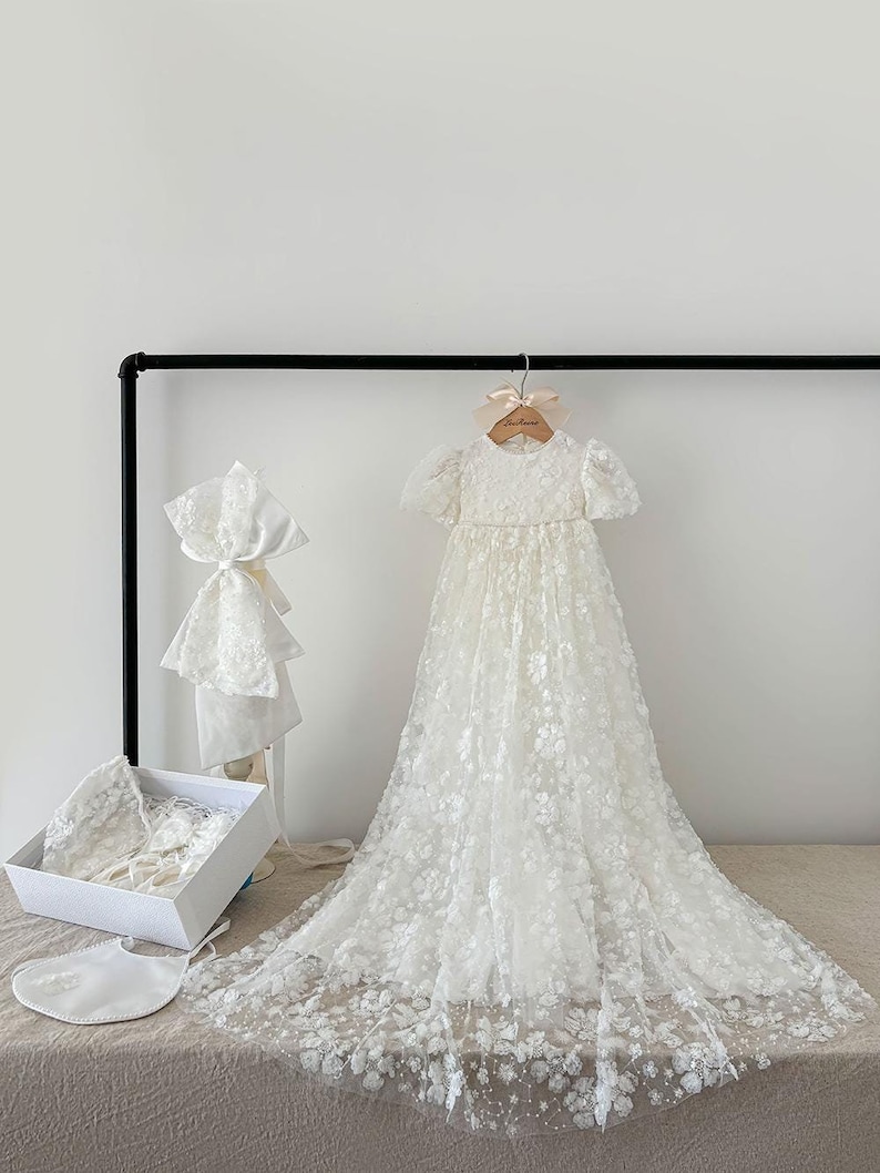 May include: A white christening gown with a lace overlay and a matching bonnet. The gown is hanging on a wooden hanger with a ribbon that says "Sweet One". The gown is displayed with a white box containing a matching bib and a white satin blanket.