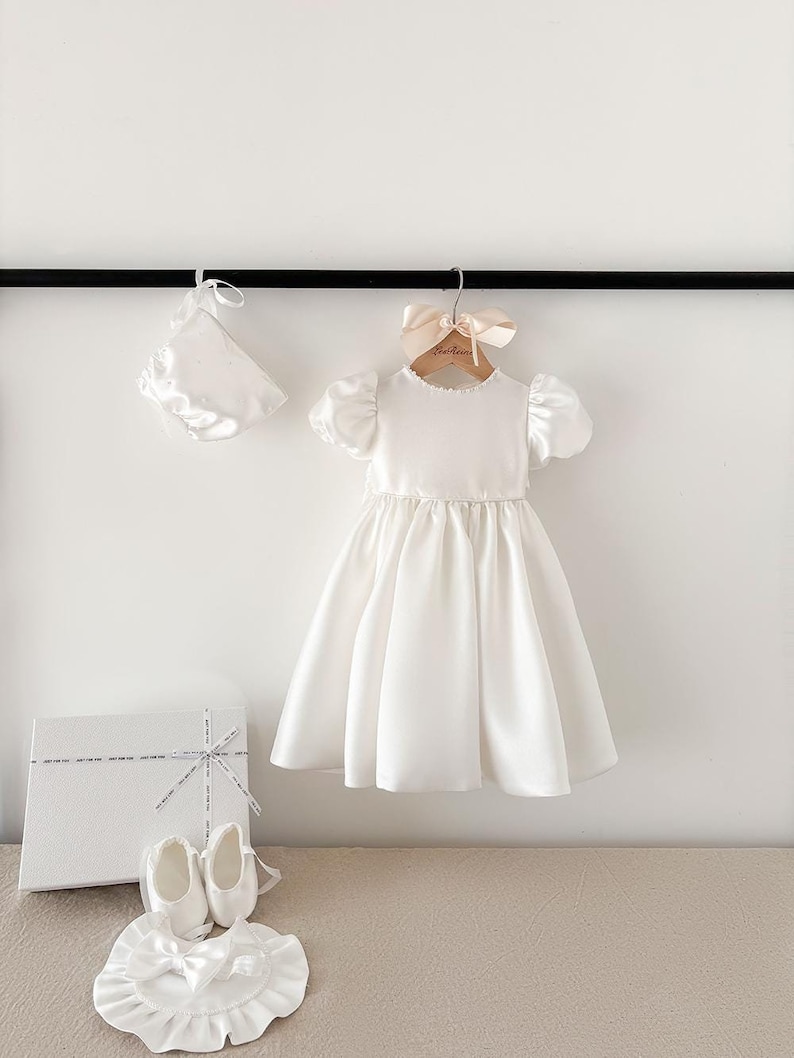 Pode incluir: Vestido de beb&eacute; de cetim branco com mangas bufantes, um gorro de cetim branco e sapatos de cetim branco. O vestido est&aacute; pendurado em um cabide de madeira com uma fita rosa. Os sapatos est&atilde;o em uma caixa branca com uma fita branca.