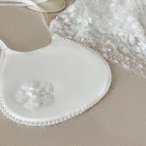 May include: A white satin bib with a pearl trim and a white flower embellishment. The bib is perfect for a special occasion.