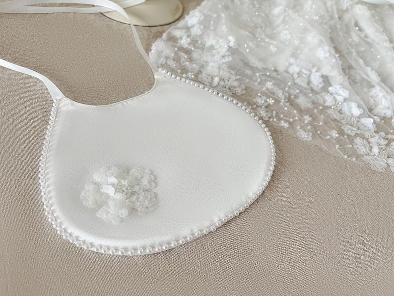 May include: A white satin bib with a pearl trim and a white flower embellishment. The bib is perfect for a special occasion.