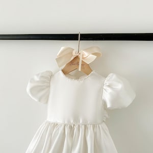 Pode incluir: Um vestido branco de cetim com mangas bufantes e uma borda de p&eacute;rolas ao redor do decote. O vestido est&aacute; pendurado em um cabide de madeira com um la&ccedil;o e o texto "Les R&ecirc;ve" impresso nele.