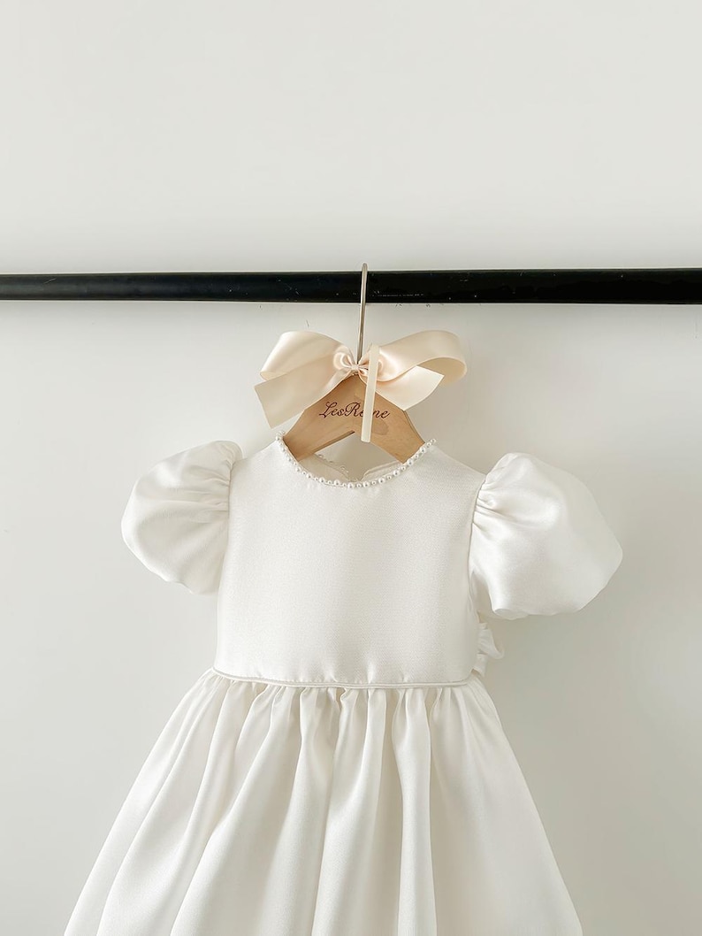Pode incluir: Um vestido branco de cetim com mangas bufantes e uma borda de p&eacute;rolas ao redor do decote. O vestido est&aacute; pendurado em um cabide de madeira com um la&ccedil;o e o texto "Les R&ecirc;ve" impresso nele.