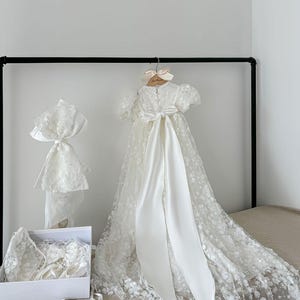 May include: White christening gown with a lace overlay and a satin sash. The gown is displayed on a hanger with a matching bonnet, shoes, and bib.