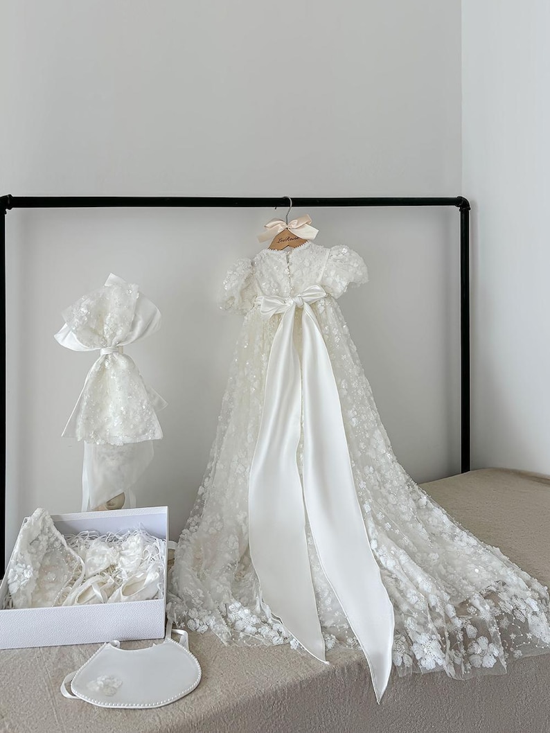May include: White christening gown with a lace overlay and a satin sash. The gown is displayed on a hanger with a matching bonnet, shoes, and bib.