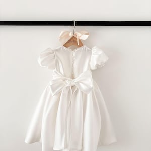 Pode incluir: Um vestido branco de cetim com um grande la&ccedil;o nas costas. O vestido tem mangas curtas bufantes e um decote redondo. O vestido est&aacute; pendurado em um cabide de madeira com uma fita rosa claro.