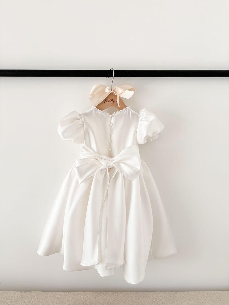 Pode incluir: Um vestido branco de cetim com um grande la&ccedil;o nas costas. O vestido tem mangas curtas bufantes e um decote redondo. O vestido est&aacute; pendurado em um cabide de madeira com uma fita rosa claro.