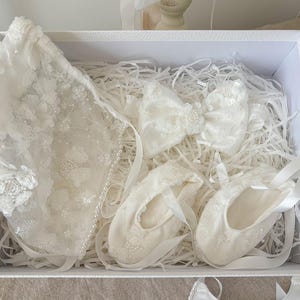May include: A white gift box filled with white lace baby clothes and accessories. The box contains a white lace bonnet, a white lace bow, and two pairs of white lace baby shoes.