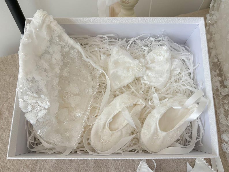 May include: A white gift box filled with white lace baby clothes and accessories. The box contains a white lace bonnet, a white lace bow, and two pairs of white lace baby shoes.