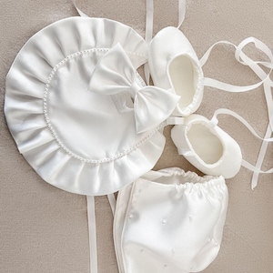 Pode incluir: Um gorro, um babador e sapatos de beb&eacute; em cetim branco com detalhes de p&eacute;rolas. O gorro tem um babado franzido ao redor da borda. O babador tem um la&ccedil;o no centro. Os sapatos t&ecirc;m um design simples.