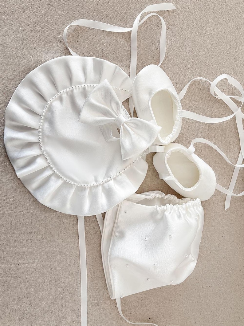 Pode incluir: Um gorro, um babador e sapatos de beb&eacute; em cetim branco com detalhes de p&eacute;rolas. O gorro tem um babado franzido ao redor da borda. O babador tem um la&ccedil;o no centro. Os sapatos t&ecirc;m um design simples.