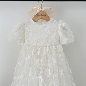 May include: A white lace dress with a beaded neckline and puff sleeves. The dress is hanging on a wooden hanger with the text "Les Reine" printed on it.