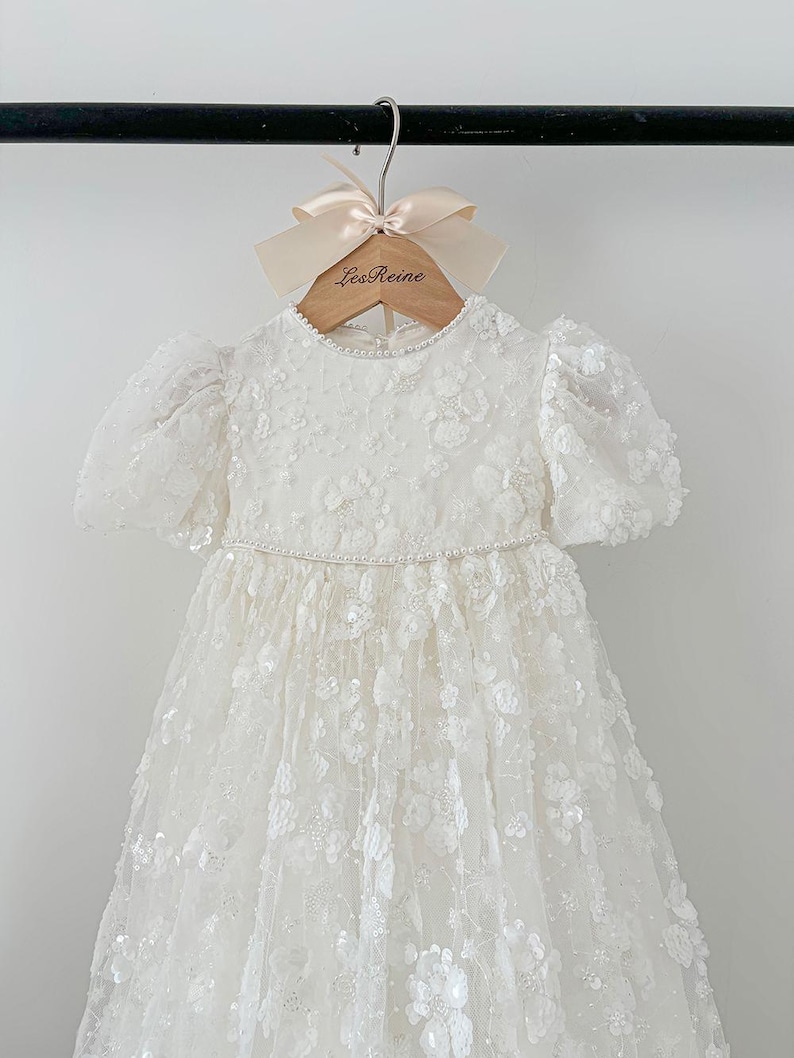 May include: A white lace dress with a beaded neckline and puff sleeves. The dress is hanging on a wooden hanger with the text "Les Reine" printed on it.