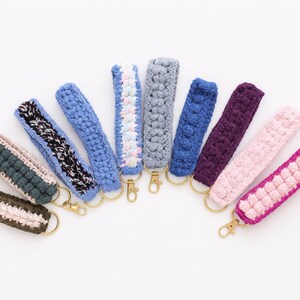 May include: A collection of handmade crochet wrist keychains in various colors, including blue, purple, pink, and gray. Each keychain features a gold-colored clasp and a unique textured pattern. The keychains are arranged on a white background.