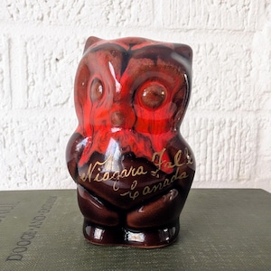 May include: A ceramic owl figurine with a glossy finish. The owl is primarily brown with red accents and has the words "Niagara Falls Canada" in gold script. The figurine is displayed on a green book.