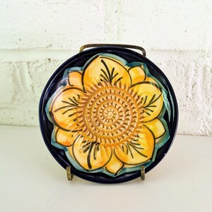 May include: A round ceramic plate featuring a central yellow sunflower design with dark brown outlines and a textured center, set against a deep navy blue background. The plate is displayed upright on a small, thin, dark metal wire stand.