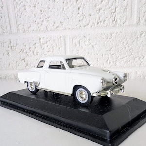 Könnte beinhalten: Ein weißes Modellauto im Coupé-Stil mit zwei Türen, chromfarbenen Stoßstangen und silbernen Felgen. Das Miniaturfahrzeug ist fest auf einem rechteckigen, schwarzen Kunststoff-Sockel mit einer glatten, erhöhten Oberfläche montiert.