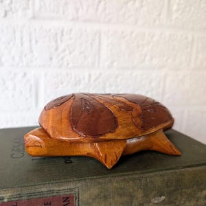 May include: A wooden turtle-shaped box with a hinged lid. The box is made of light and dark brown wood, with carved leaf details on the lid. The turtle box is resting on a vintage book.