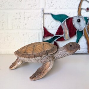 May include: A detailed, wooden turtle figurine with a textured, brown shell and light brown limbs. The turtle is positioned on a white surface, with a stained glass fish in the background. The figurine has a realistic appearance.
