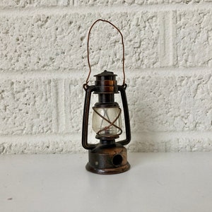 May include: A vintage metal pencil sharpener shaped like a lantern. The sharpener is brown and has a clear plastic window. It has a wire handle and a hole for sharpening pencils.