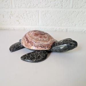 May include: A decorative stone turtle figurine. The turtle's shell is a mix of brown and pink hues, while the head, legs, and flippers are dark gray. The figurine is placed on a white surface against a white brick wall.